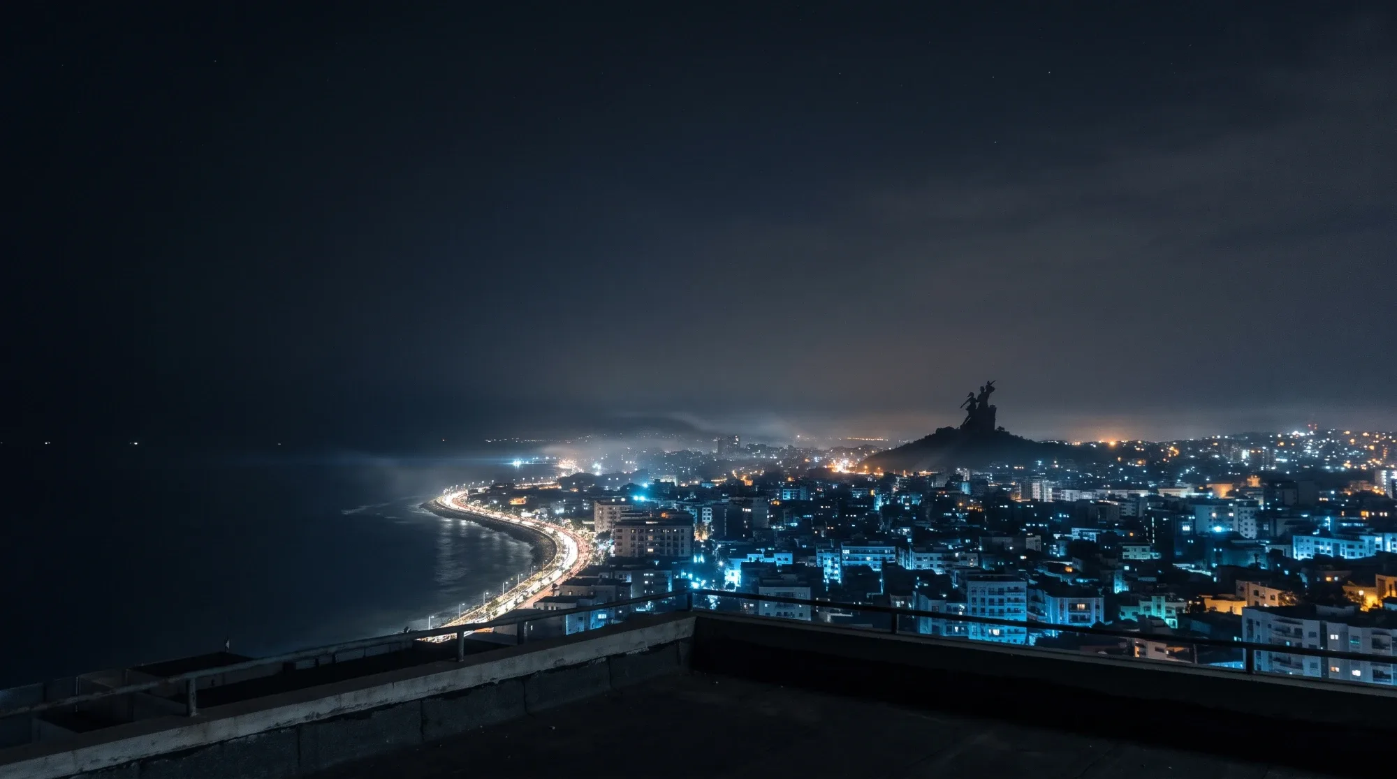 Dakar at night seen from a rooftop, the lit Corniche and the African Renaissance Monument on its hill